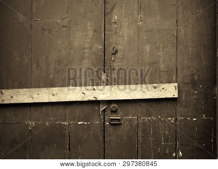 Sepia Close Up Of An Old Cracked Peeling Wooden Plank Door Barred Shut With A Piece Of Timber And Ru