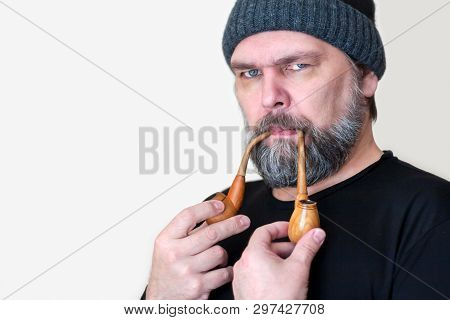 A Serious Bearded Middle-aged Man In A Knitted Cap Smokes Two Smoking Pipes.