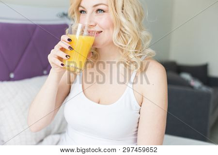 Attractive Blonde Woman Sits On The Edge Of The Bed At Home And Holds A Stocker With Orange Juice In