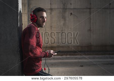 Muscular Athetic Man Wearing Sportswear, Leaning Onto A Concrete Wall, Choosing A Song On A Smart Ph