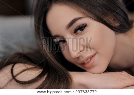 Beautiful Brunette Girl With Brown Eyes With Bare Shoulders Lying On The Bed. Face Close Up. Asian A