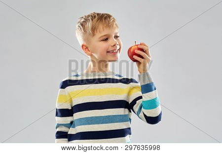 food, diet and children concept - smiling boy in striped pullover eating red apple over grey background
