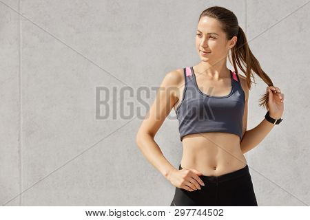 Studio Portrait Of Young Beautiful Sporty Woman, Wearing Gray Sports Top And Black Leggins, Touches 