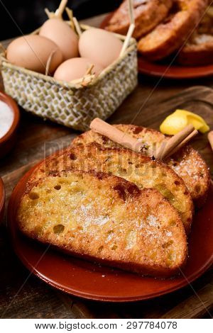 an earthenware plate with some torrijas, typical spanish dessert eaten in Lent and Easter, and some of the ingredients to prepare it, such as eggs, sugar or lemon peel, on a wooden table