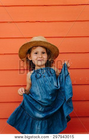 Happy Child Girl In Summer Hat And A Beautiful Blue Dress Against Orange Wall. Lifting Her Skirt In 