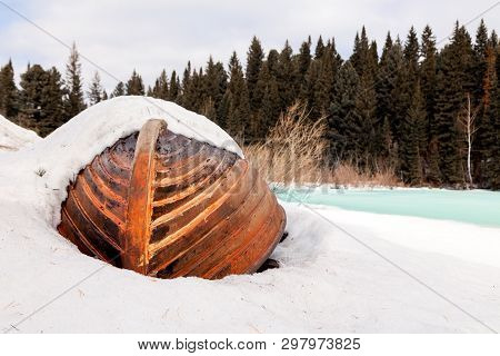 Old Wooden Destroyed Flooded Fishing Boat Lie In Snow On Shore Of Ice-covered Frozen River In Winter