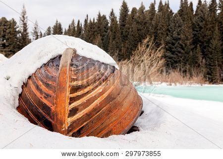 Old Wooden Destroyed Flooded Fishing Boat Lie In Snow On Shore Of Ice-covered Frozen River In Winter