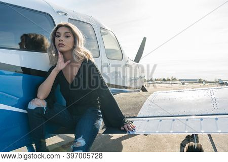A gorgeous young blonde model poses outdoors with a single engine aircraft at a local airport
