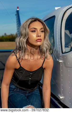 A gorgeous young blonde model poses outdoors with a single engine aircraft at a local airport