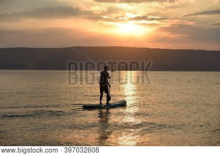 Man Sail On A Sup Board In A Large Lake During Sunrise. Stand Up Paddle Boarding - Active Recreation