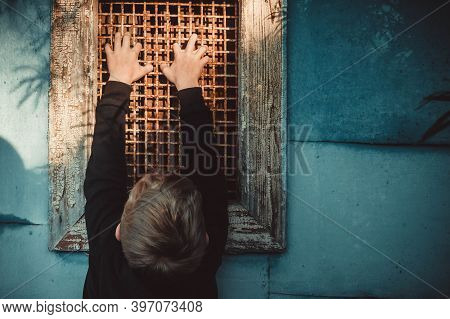 Window With Bars. A Hand Gripped An Old Metal Grate. The Boy Is Holding On To The Bars. The Child Cl