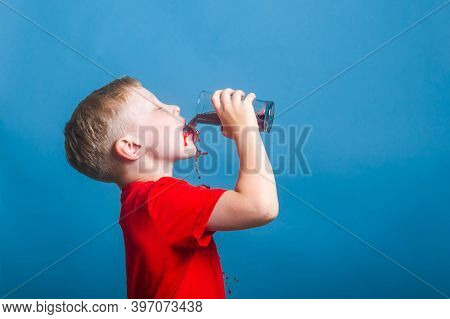 A Blond European Boy Of 6 Years Old Drinks Juice From A Glass. A Child In A Red T-shirt Splashes And