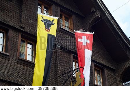 Banners Of The Canton Uri And Switzerland Hanging In Front Of A Typical Wooden Chalet In Andermatt, 
