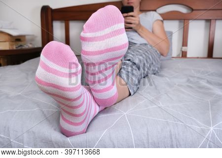 Crossed Feet In Cozy Cotton Striped Pink And White Socks Of A Woman Sitting On A Bed And Relaxing Wh