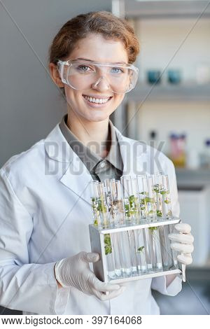 Vertical Portrait Of Young Female Scientist Smiling T Camera While Holding Test Tubes With Plant Sam