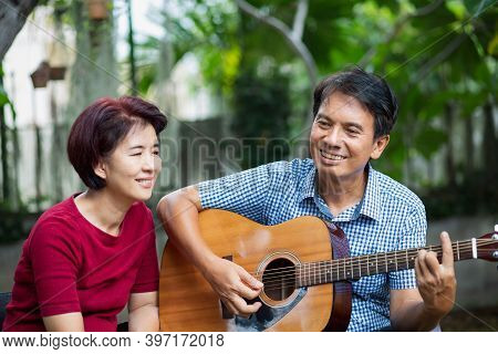 Middle Aged  Couple Playing Guitar While Relax Sitting On Bench In Backyard.