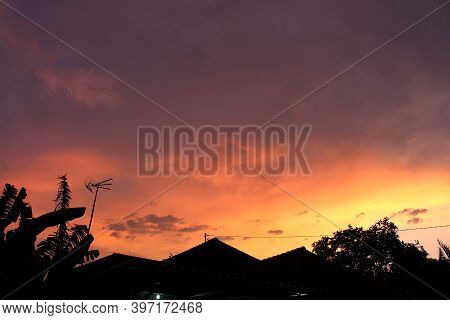 The Evening Sky Is Blue And Orange. There Are Silhouettes Of Roofs Of Houses And Trees