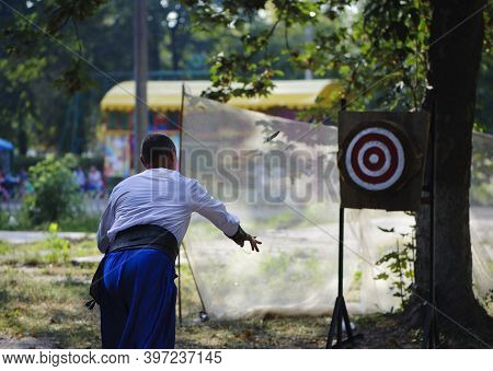 A Man Throws Knives At A Wooded Target. White Shirt And Blue Pants On A Young Guy. Competitions, One