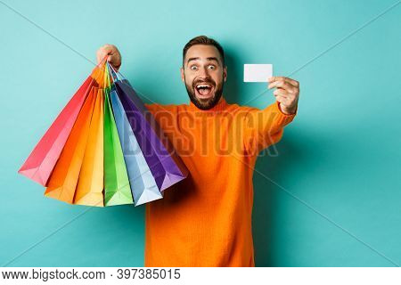 Happy Aduly Man Showing Credit Card And Shopping Bags, Standing Against Turquoise Background