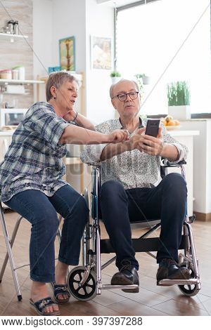 Disabled Senior Man In Wheelchair And His Wife Are Browsing On Internet Using Smartphone. Paralysed 