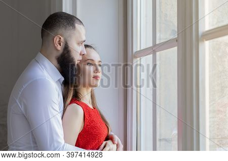 Sad Young Couple Looking In The Window On Sunny Day. Man And Woman Hugging At Home After A Quarrel, 