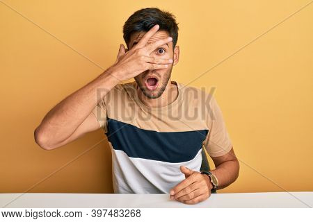 Young handsome man wearing casual clothes sitting on the table peeking in shock covering face and eyes with hand, looking through fingers afraid 
