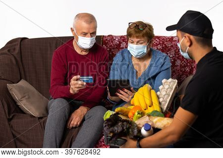 Senior Couple In Medical Masks Orders By Courier And Paid By Credit Card. Pandemic, Online Concept.