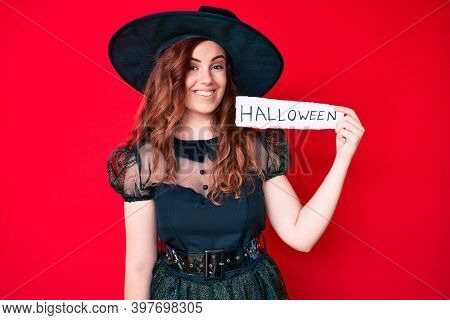 Young beautiful woman wearing witch costume holding paper with halloween message looking positive and happy standing and smiling with a confident smile showing teeth 