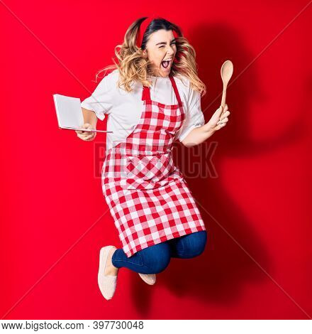 Young beautiful blonde cook woman wearing apron smiling happy. Jumping with smile on face holding recipe book and spoon over isolated red background