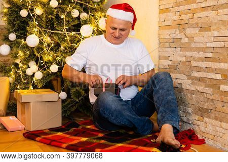 Grandfather Is Sitting Near Cristmas Tree In Santa Clauss Hat At Night At Home.