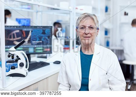 Senior Researcher Sits At Her Workplace In Laboratory Looking At Camera. Elderly Scientist Wearing L