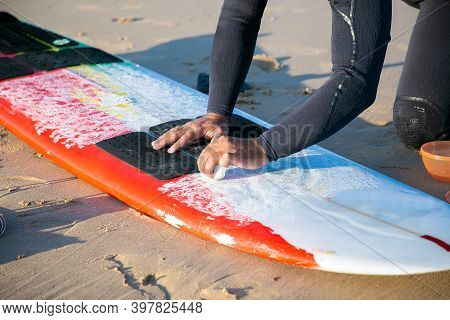 Hands Of Male Surfer In Wetsuit Waxing Surfboard On Sand On Ocean Beach. Cropped Shot. Surfing And A