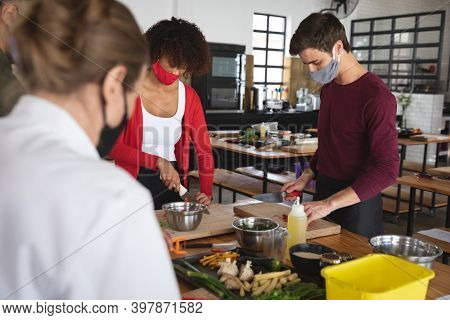 Female chef and divserse group wearing face masks in kitchen. trainee chefs standing around a table listening to female chef. hygiene in professional kitchen during coronavirus covid 19 pandemic.