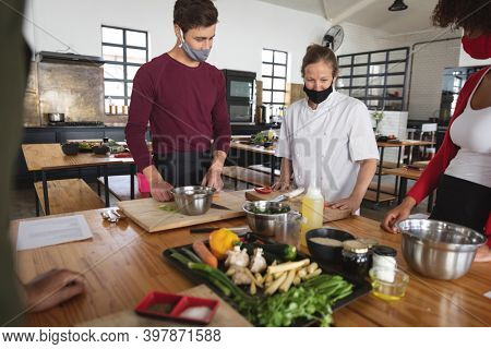 Female chef and divserse group wearing face masks in kitchen. trainee chefs standing around a table listening to female chef. hygiene in professional kitchen during coronavirus covid 19 pandemic.