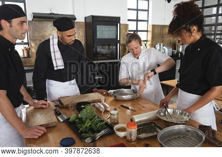 Female chef and divserse group in kitchen. trainee chefs standing around a table listening to instructions from female chef.