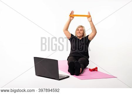 Woman With Elastic Band Sitting On The Mat In Front Of The Laptop. Pandemic On White Background.