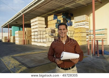 Portrait of happy middle aged man with clipboard standing in front of timber factory