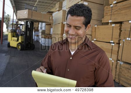 Happy middle aged man writing on notepad with worker working in forklift at warehouse