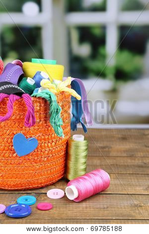 Round orange box full of multicoloured thread and everything necessary for sewing on a brown wooden table on window background