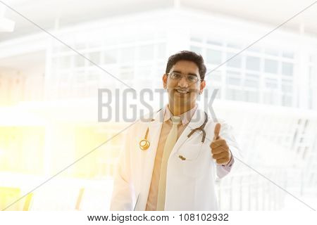 Portrait of a smiling Asian Indian male medical doctor in uniform thumb up, standing outside hospital building block, beautiful golden sunlight at background.