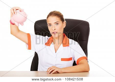 Young female doctor or nurse sitting behind the desk and holding a piggybank.