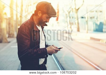 Half length portrait of bearded male with retro style using cell telephone while standing outdoors