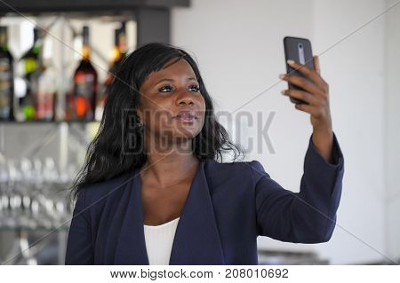 young attractive and happy black afro american woman in casual elegant clothes taking selfie portrait photo with mobile phone camera smiling cheerful and positive sitting at restaurant bar
