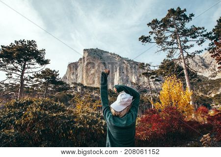 Back view of traveler hipster girl wearing cap and sweater standing alone in autumn woods and looking at mountain hill. Cold weather, fall colors. Wanderlust concept.