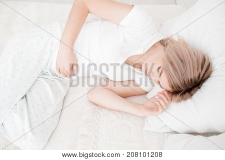 Blonde, young girl in white clothes lies on a bed with a pain in the stomach on a white background