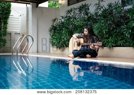 young asia woman hold guitar classic in her hand and sitting beside pool around her has peaceful green nature environment. image for travel person music instrument place nature portrait concept