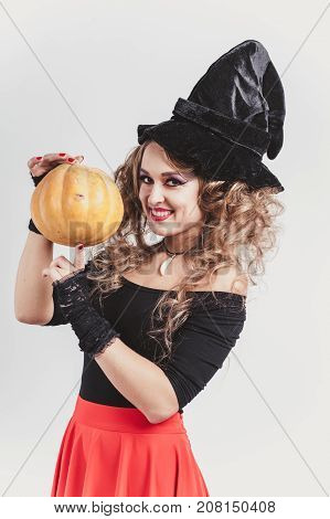 Portrait of woman holding in hands orange pumpkin near face isolated on gray background. Wearing black witch hat, blouse and red skirt. Halloween concept. Vertical shot