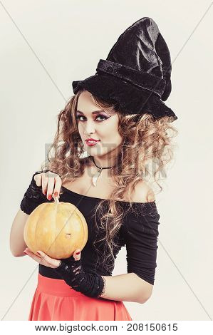Portrait of woman holding in hands orange pumpkin isolated on gray background. Wearing black witch hat, blouse and red skirt. Vertical shot
