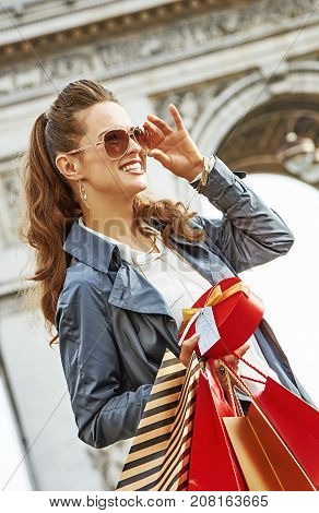 Stylish Christmas in Paris. smiling elegant woman in trench coat with shopping bags and Christmas present near Arc de Triomphe in Paris France looking into the distance