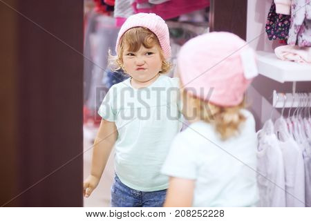 Adorable toddler girl in baby apparel store buying warm autumn clothes fiting knitted hat disappointed dace.